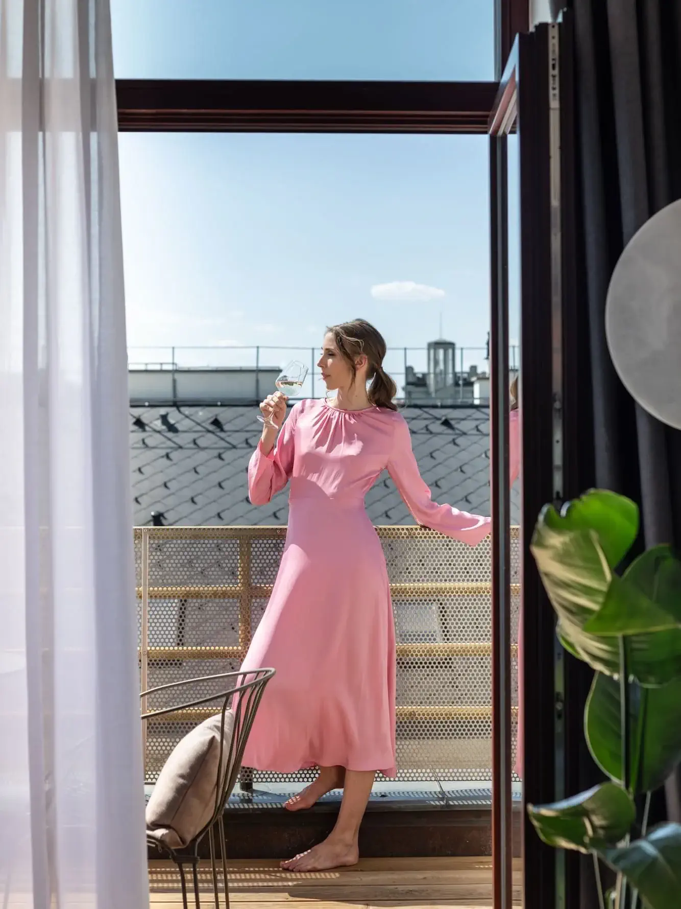 Eine Frau in einem langen rosa Kleid steht barfuß auf einem Balkon, hält ein Weinglas in der Hand und schaut an einem sonnigen Tag nach draußen.